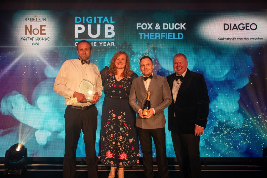 Three men and a woman in evening attire. The man on the left holds a glass award and the man second from the right holds a bottle of champagne. Above them the background says "Greene King Night of Excellence 2024, Digital Pub of the Year, Fox and Duck, Therfield."
