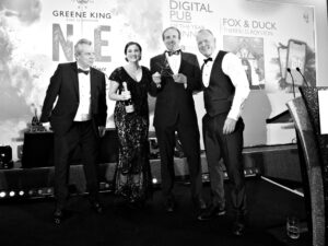 A woman and three men in evening dress. The man second from the right is holding a star-shaped glass award and the woman, who is standing second from the left, is holding a bottle of champagne. Behind them it says "Digital pub of the year winner, Fox and Duck, Therfield, Royston".